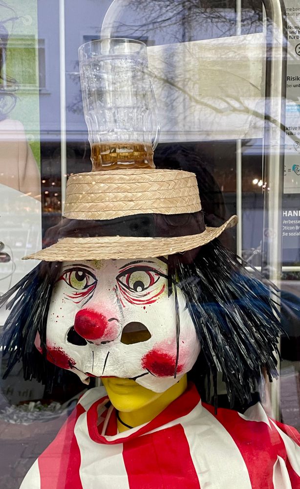 Auf dem Bild sieht man eine bunt gestaltete Schaufensterfigur mit einer clownartigen Maske. Die Maske hat einen roten Nasenknopf, stark geschminkte Wangen und dunkle, zerzauste „Haare“. Die Figur trägt einen Strohhut, auf dem ein umgedrehtes Bierglas steht. Das Outfit ist rot-weiß gestreift, ähnlich einem Narrenkostüm.