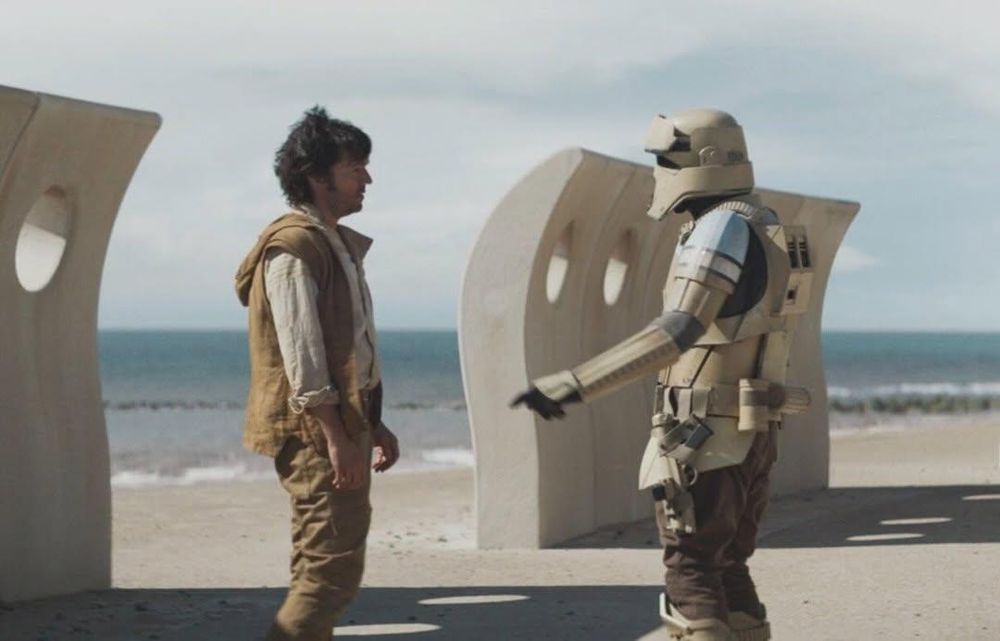 A towering enforcer droid lifts and pins a man by the throat against a concrete wall along a desolate beach. The man dangles helplessly, wide-eyed, as the machine’s grip tightens. A stark metaphor for where we are in this administration—people feeling throttled and suspended in midair, powerless against cold mechanical authority.