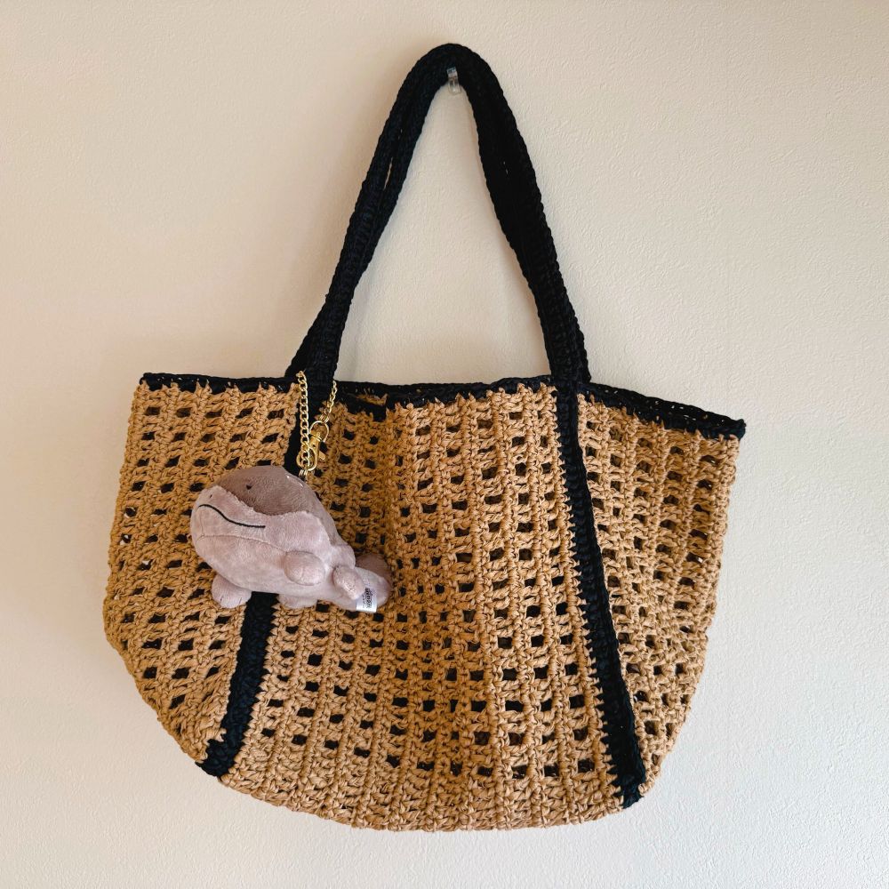 かぎ針編みのトートバッグ（ライトブラウンに黒のライン、ぬいぐるみのチャームつき）が白い壁にかけてある。