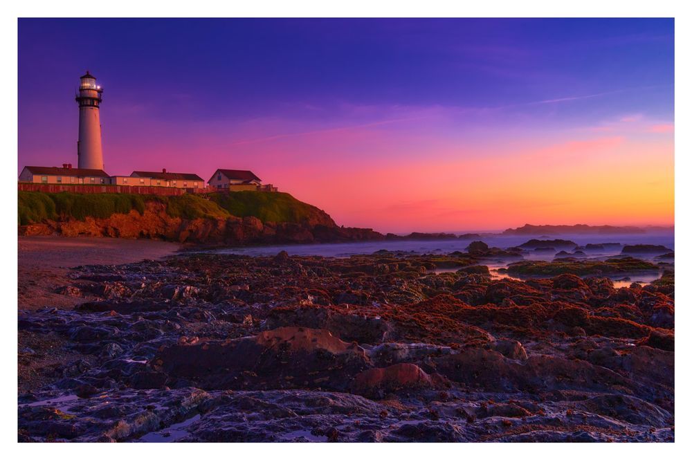 A picturesque lighthouse perched on a rocky coastline at sunset, with vibrant purple and orange skies. Nearby buildings are situated on a grassy cliff, while the tide reveals various rock formations along the shore.