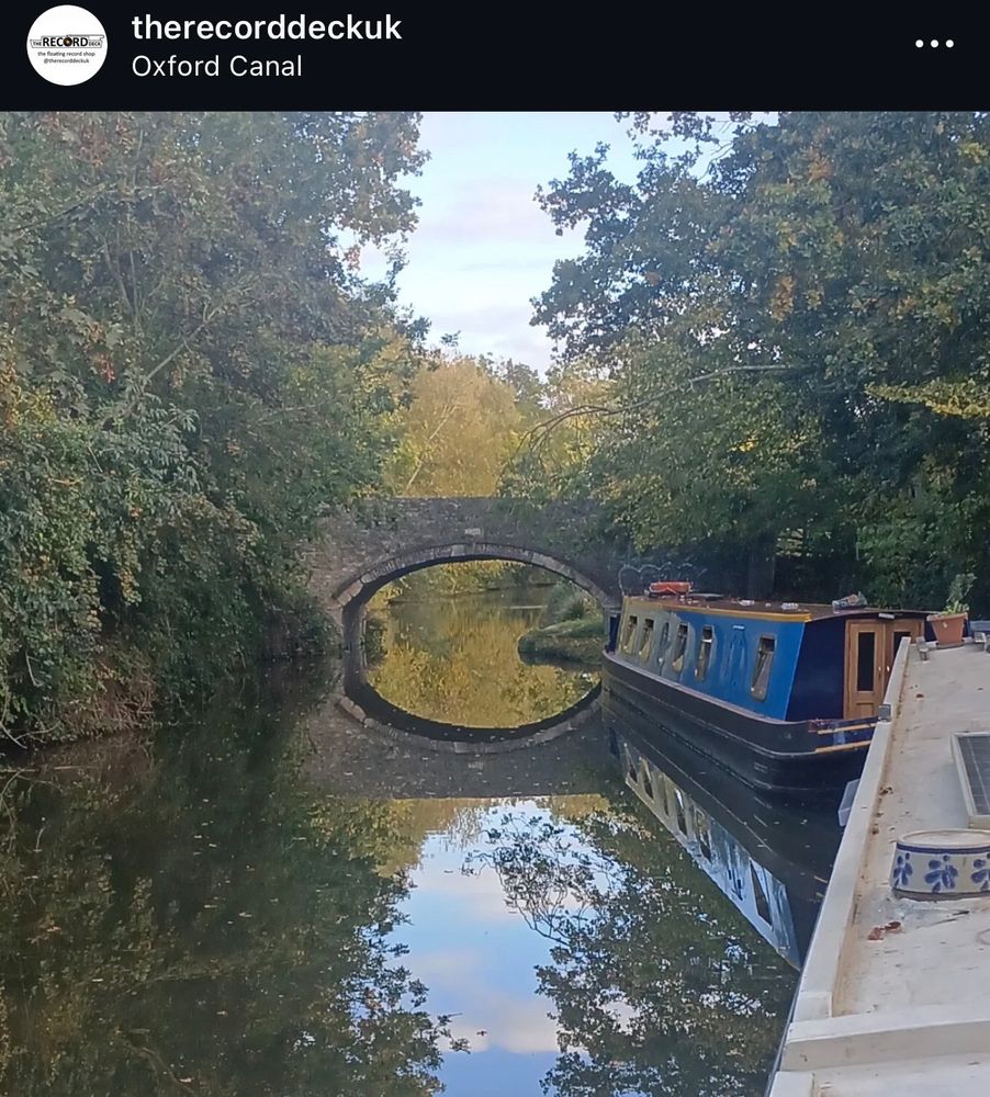 A photo from a barge on the Oxford Canal taken by the owner of The Record Deck, which is a floating record shop in the UK. It shows a bridge reflected in the smooth waters that makes it look like some kind of weird interdimensional portal to another world. Which it probably is. 