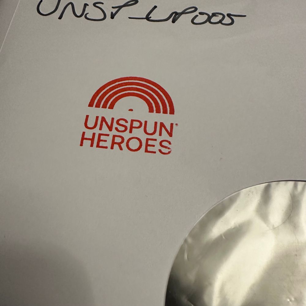 An image of a white inner sleeve featuring a red logo that reads "UNSPUN HEROES" with an arc design resembling a record being removed from a sleeve. The top portion includes handwritten text that is the catalogue number UNSP-LP005. A circular area on the right suggests this inner sleeve houses a vinyl record, which is correct. It’s a test pressing.