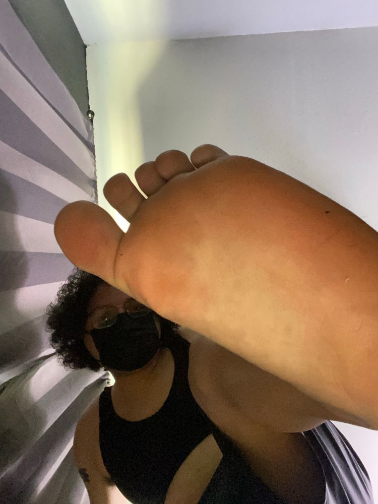Bea lifts her left foot up in the air. This picture is a closer shot of Bea. She’s wearing a black bra, black shorts, glasses, & a black mask. 
