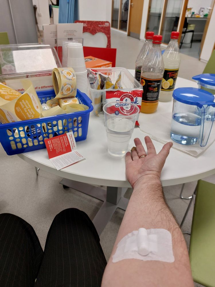 Outsterched arm with plaster on inside of elbow in front of table with crisps, biscuits, cordials and jars of water