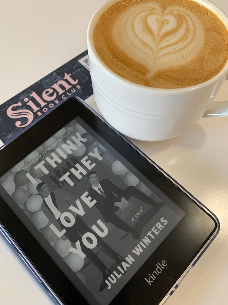 I Think They Love You by Julian Winters on ereader screen with silent book club bookmark next to cup of coffee with foam heart. 