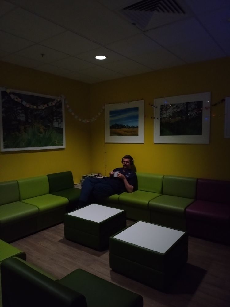 Me, in navy blue scrubs, at chaise in the corner of the staff room ~03.00 on night duty. 
I have a cup of tea, I look displeased.
The walls of the staff room and the box seating are various colours, similar to vomitus aspirate.