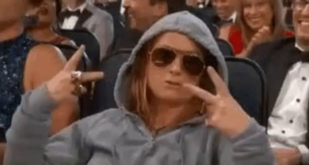 a woman wearing sunglasses and a hoodie is sitting in a crowd of people and making a peace sign .