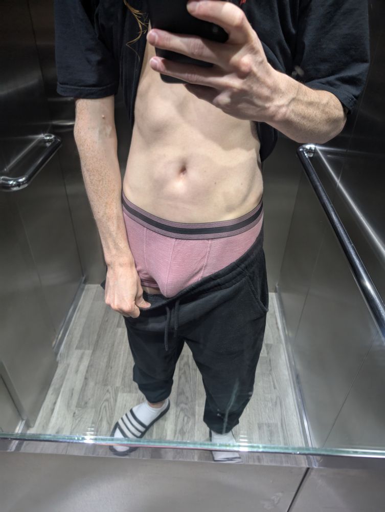 The photo is taken in the mirror of an elevator. George has pulled his tshirt up, and his joggers down, revealing his tummy and pink boxers. His cock is soft yet a large bulge is visible.