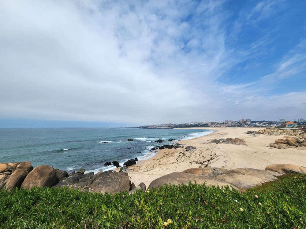 A bright clear day over a sandy beach with greenery in the foreground and Porto city in the distance. Clouds gather off the coast
