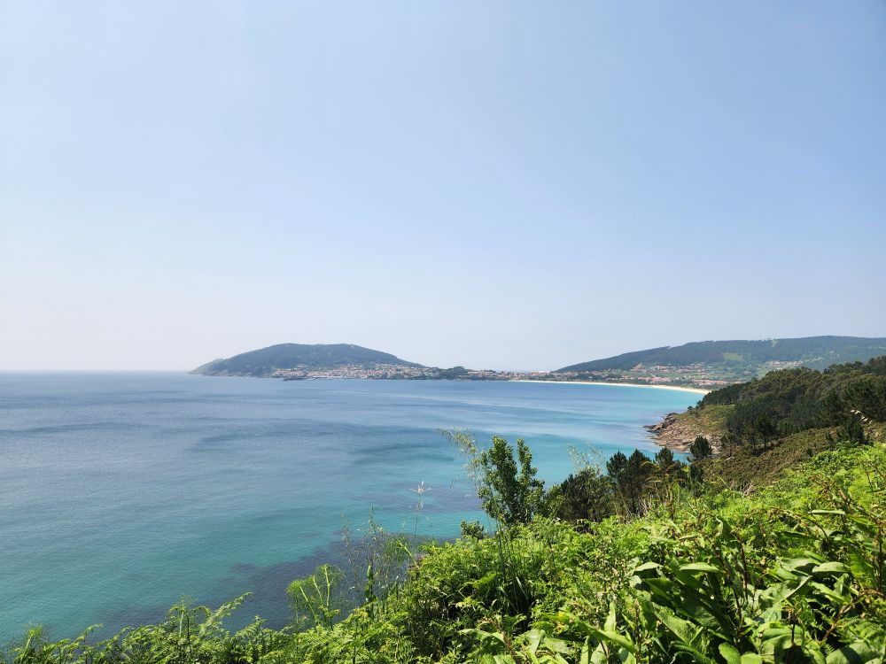 A view of finesterre, the terminus of the camino trail, can be seen in the far off distance across the ocean and sandy beach. Ferns and trees cover the forefront