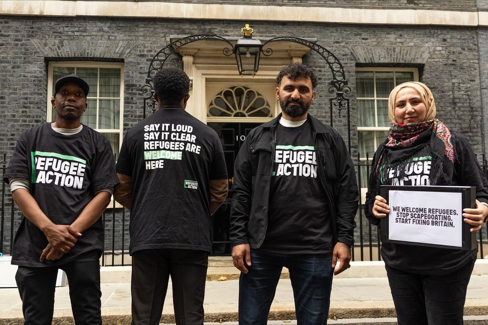 Four activists wearing Refugee Action t-shirts stand in front of 10 Downing Street, holding signs that support refugee rights in the UK.