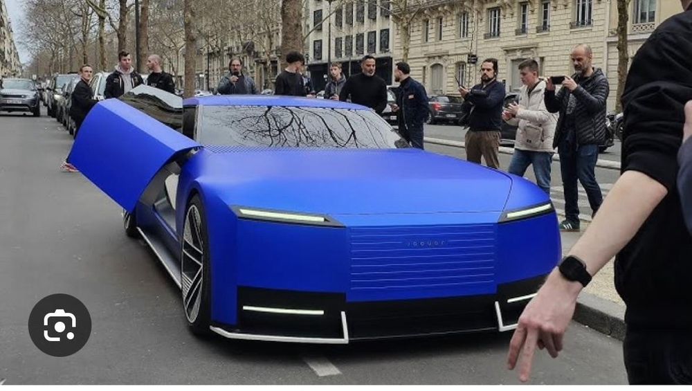 Hard to describe, the new Jaguar Type00 in aquamarine blue on a Paris street, very blue, very sleek, sort of matt, surrounded by photographers. The car is blue with black and white details.