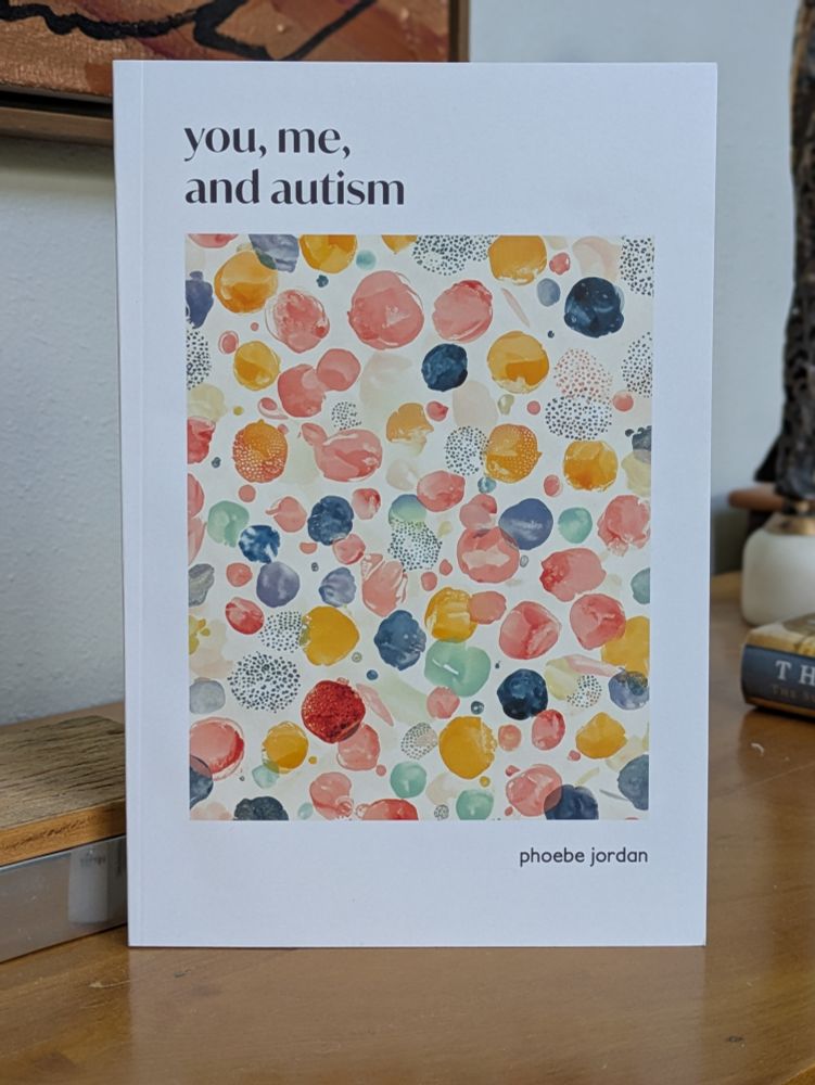 Photo of the book You, Me, and Autism by Phoebe Jordan. The book is standing upright on a wood table. The cover shows a brightly colored abstract painting of dots that reflect the diversity of autistic people.