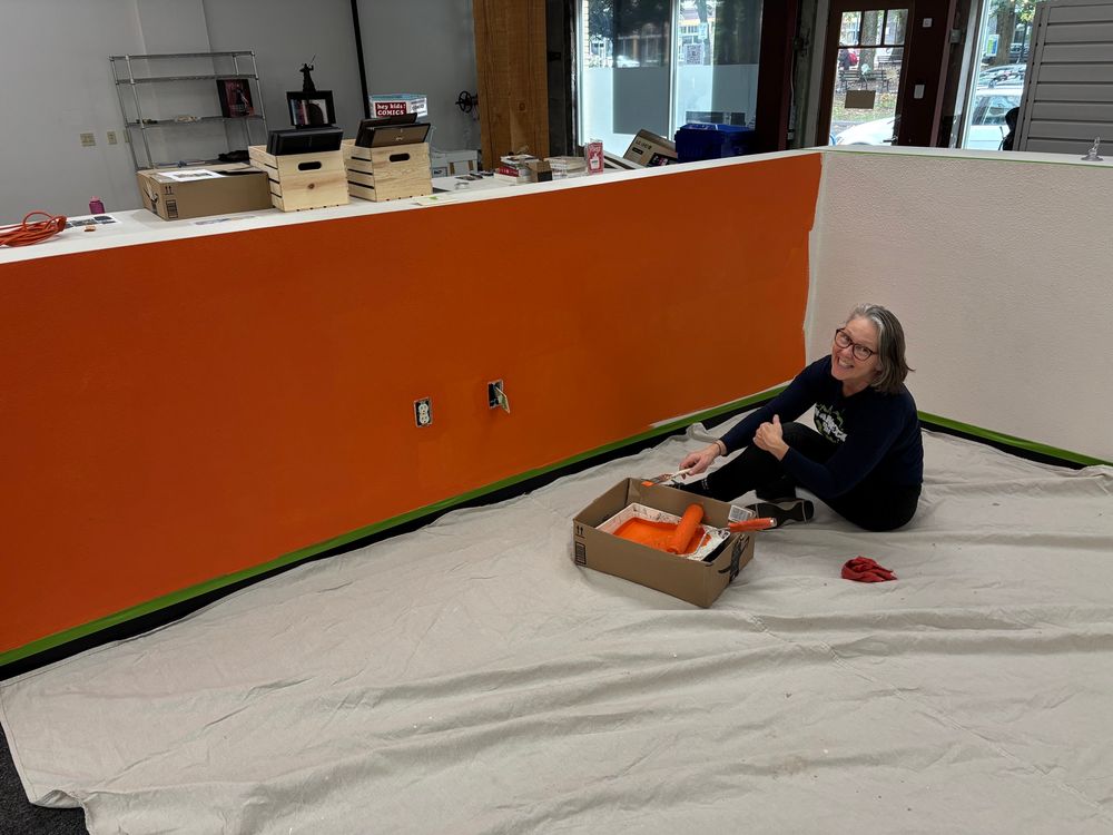 Terri is on the floor painting a wall orange. 