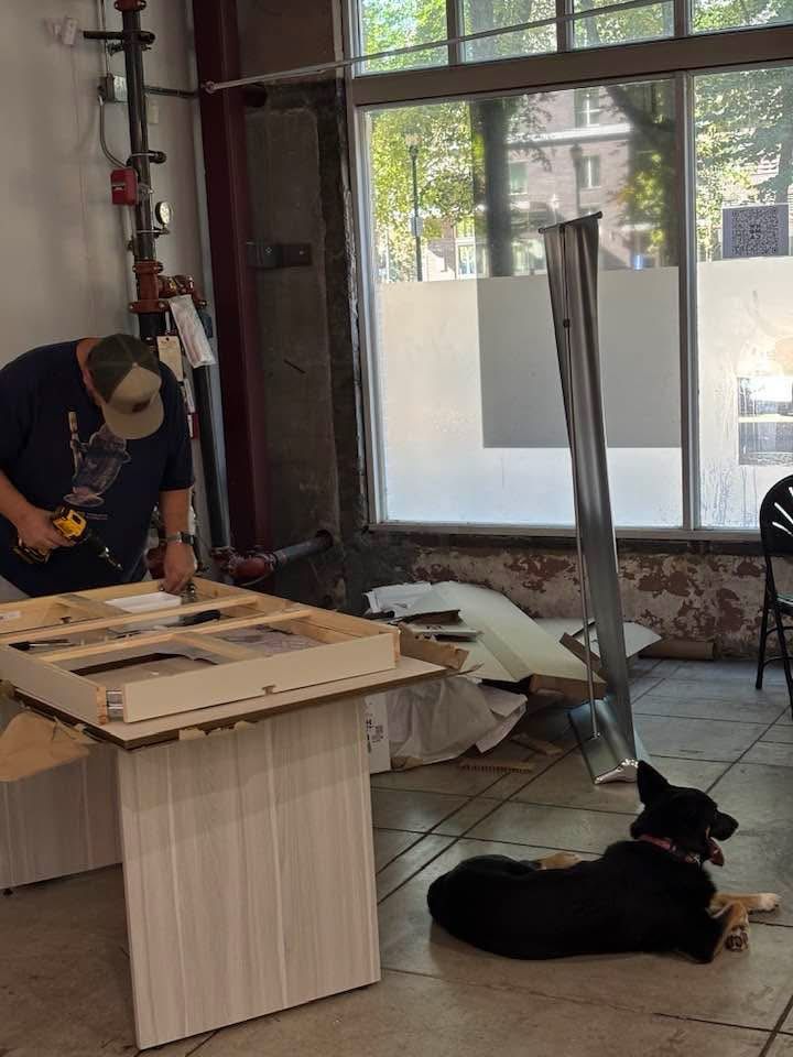 A guy puts together a table while a dog supervises. 