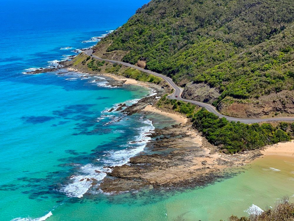 A photo of coastline called the great Ocean road in Victoria Australia. The water is blue and torquise. The waves crash on to rocky alcove and sandy beach. The conference is hosted at one of the local towns.