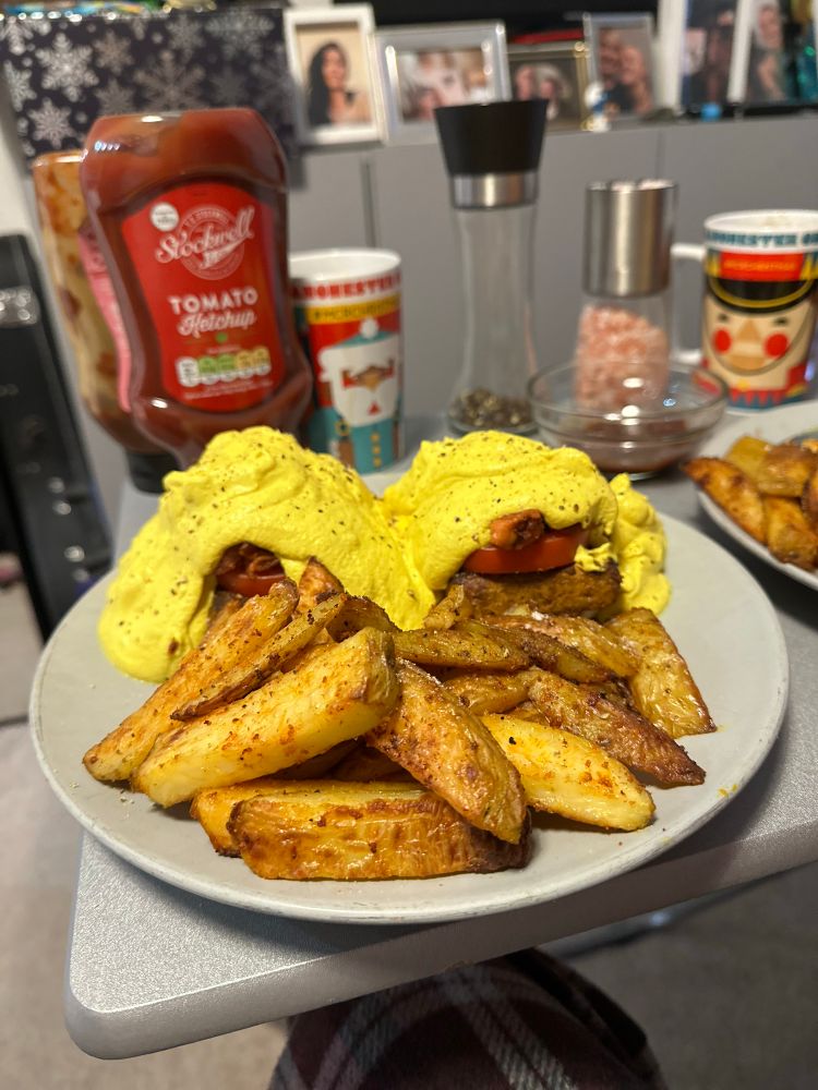 A photo of vegan eggs Benedict and home fries on a grey plate on a grey tray table in front of gray cabinets. On the tray table alongside the dish are Manchester Christmas market mugs filled with coffee, a salt shaker and a bottle of ketchup. In the background atop the cabinets are framed photos of those that are near and dear to me.