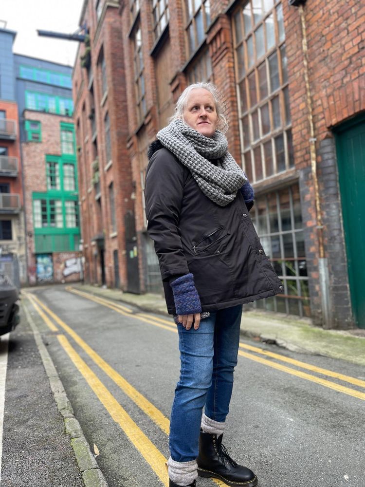 Middle-aged grey haired older person in front of old buildings in Manchester wearing winter wear that includes a black winter coat with lots of pockets, a giant grey scarf, blue jeans, grey comfy socks and doc martins