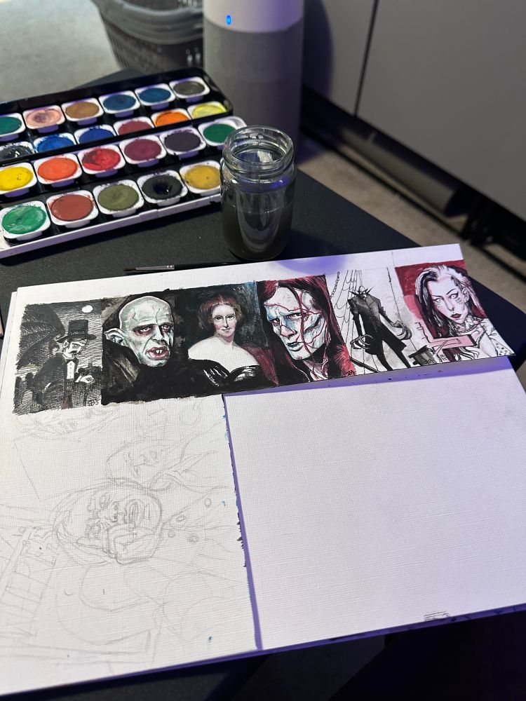 Tiny watercolour paintings on a tv dinner table plus art supplies including a jar of paint water, a watercolor set, some brushes and a pencil and eraser. The paintings are of Dracula, Klaus Kinski as Nosferatu, Mary Shelley, Jacob Elordi as Frankenstein’s Monster, Nosferatu and my beloved Friend Xy who these paintings are for.