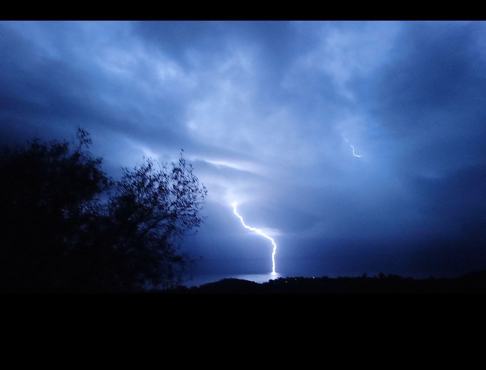 La foudre tombant sur la Mer dans un ciel bleuté magnifique rn arrière plan, on y voit au second des oliviers remués par le vent. Photo nocturne de Corse.