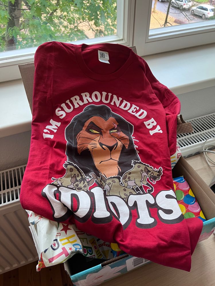 Ein Shirt, auf dem Scar aus „König der Löwen“ abgebildet ist, dazu der Schriftzug „I‘m surrounded by idiots“. Darunter ein Postkarton, aus dem Geschenkpapier guckt.