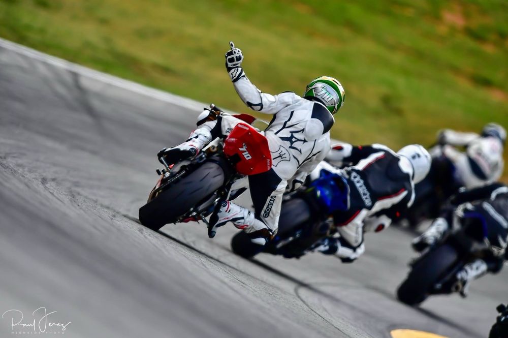 photo from behind a man on a red motorcycle in white racing leathers & a helmet giving the finger as he goes around the track