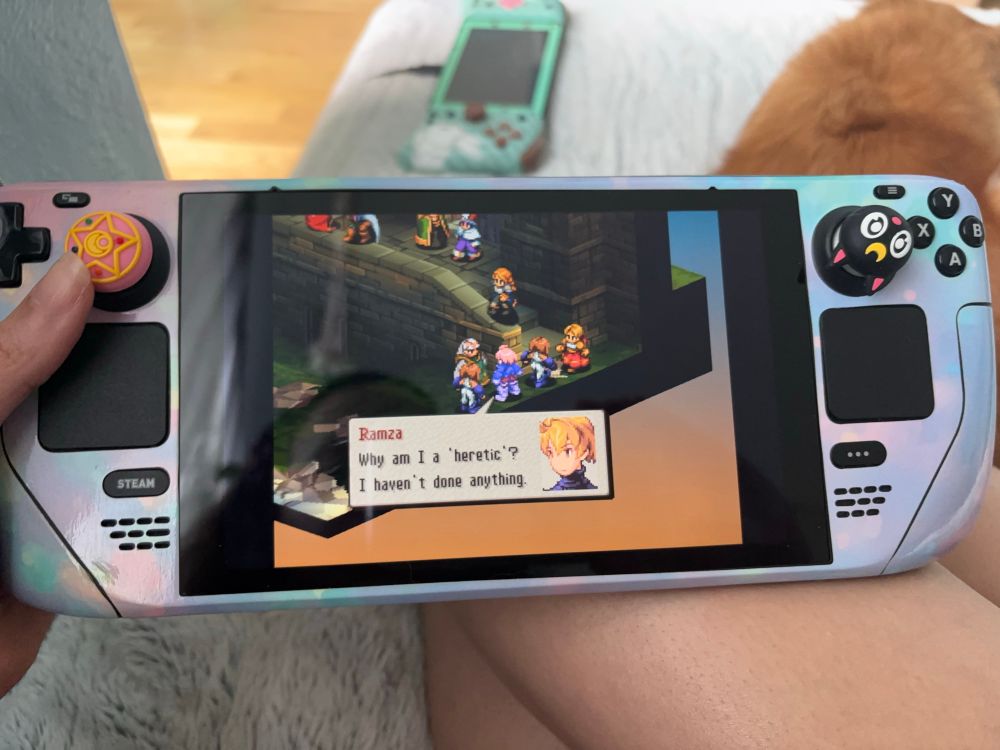 Mars holding her steam deck playing Final Fantasy Tactics. The character Ramza is speaking, he says, “Why am I a ‘heretic’? I haven’t done anything.”