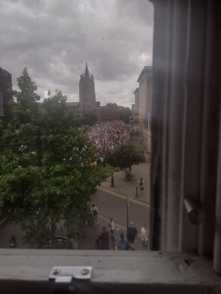 Corner of a window frame. Outside, can see a tree, an old church in the distance, and a crowd of people with pride flags