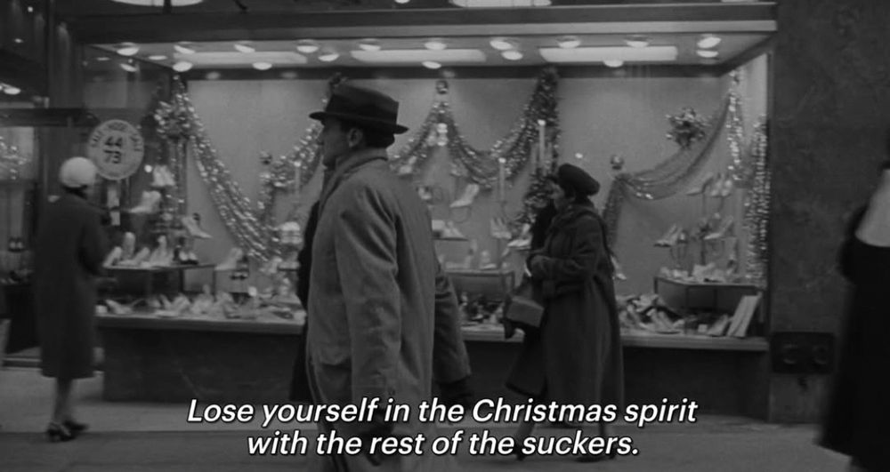 Black and white still of a film. A man in a suite walks down an xmas decorated street. The subtitles read ‘Lose yourself in the Christmas spirit with the rest of the suckers.’