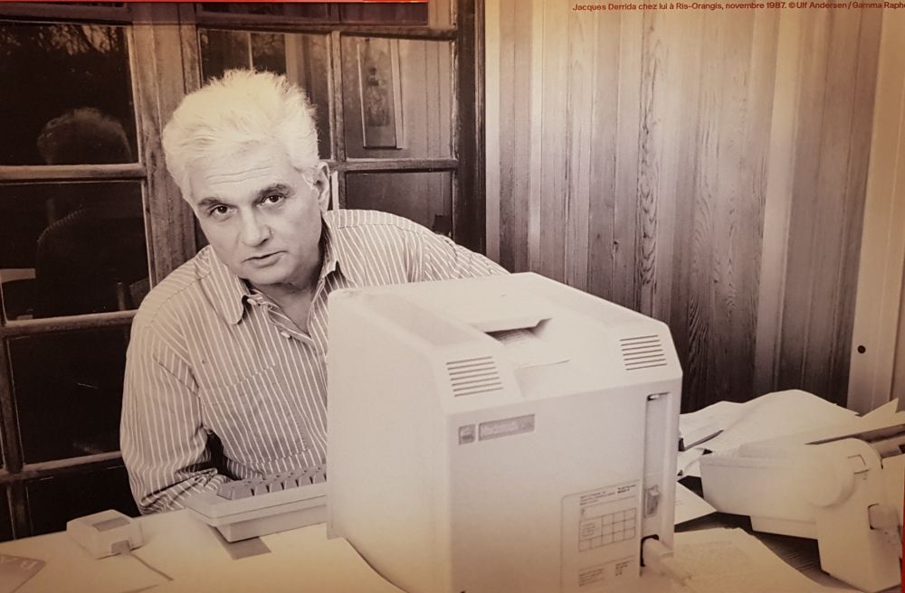Jacques Derrida guckt hinter einem sehr altmodischen Computer hervor, ein schwarz-weiss Foto von 1987