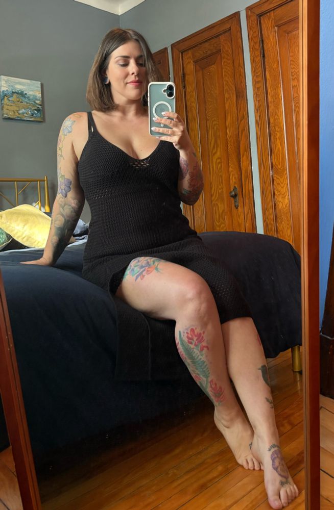 A tattooed brunette with shoulder length hair wearing a black knit dress sits on a bed, smiling.