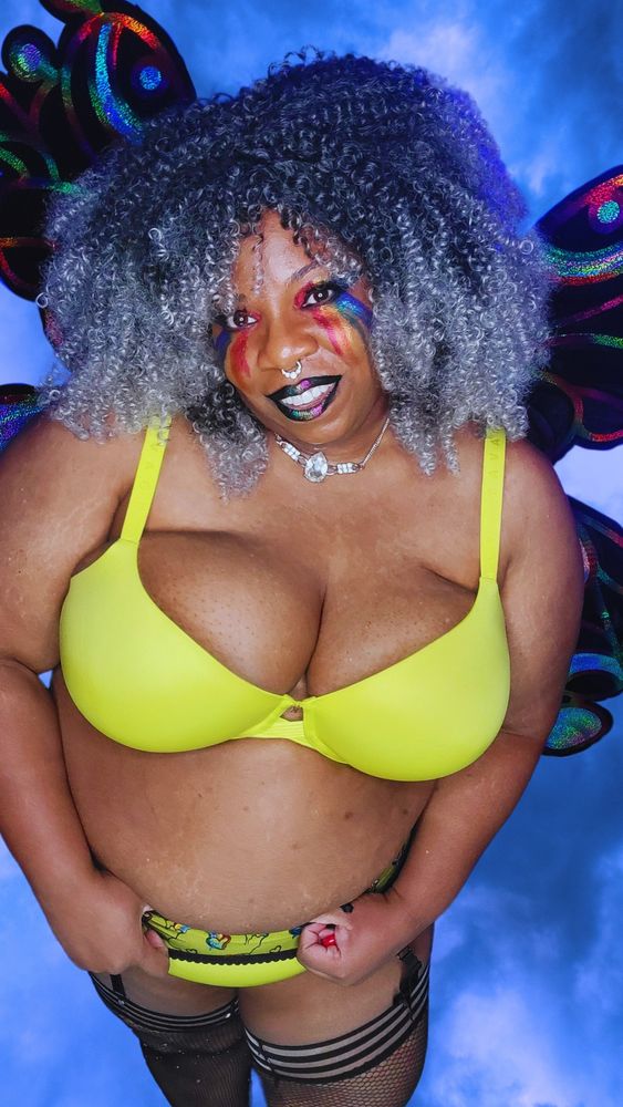 TaLynn Kel, a fat Black femme, is wearing a yellow bra, yellow underwear, a black garyer belt with rainbows on it, and black fishnet thigh highs with little jewels. She has curly black to grey ombre hair and black butterfly wings with rainbow trim. She has red, gold, and blue eyeshadow on her top lids with red, orange, yellow, green, blue, and purple streaks trailing down her face. She is wearing black lipstick with a rainbow stripes across the middle of her lips. She is standing and smiling at the camera. 