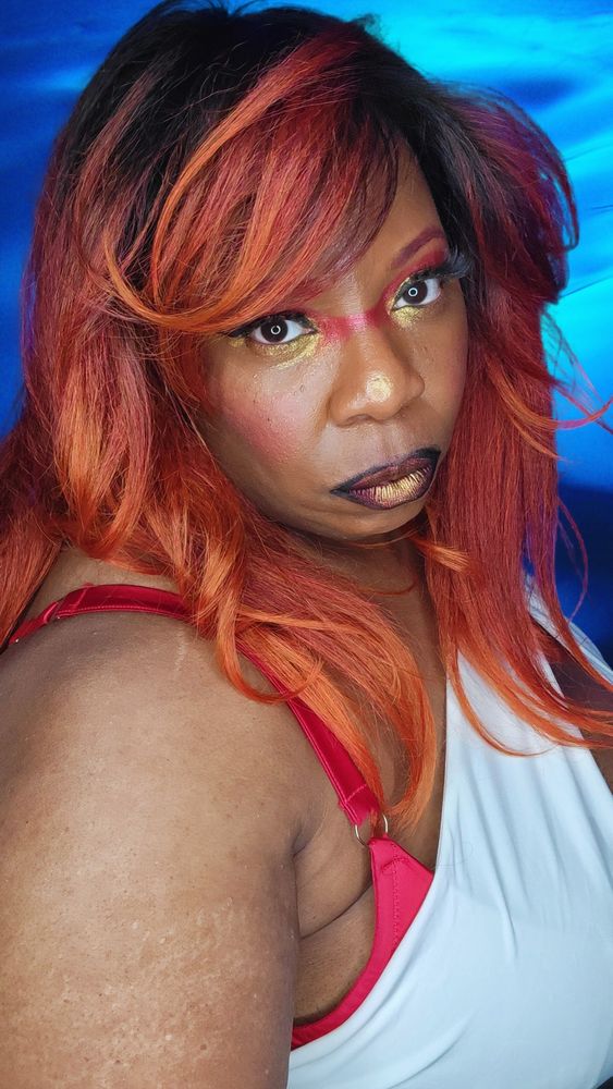 TaLynn Kel, a fat Black femme with ombre black, red, and copper hair, is wearing an asymmetrical white top with a cleavage cut-out and a red bra. She has red and gold eye makeup and black, red and gold lipstick. She is sideways with her face turned towards the camera with an intense look on her face. 
