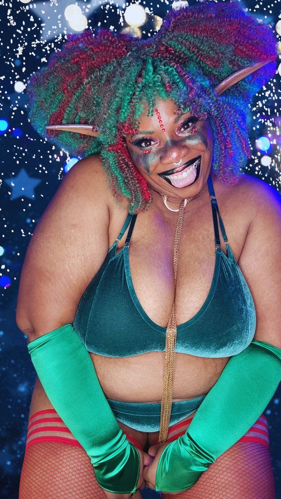 TaLynn Kel, a fat, Black woman, has curly red and green hair, long pointy ears, and red and green makeup. She's wearing a green velvet bra and panties, green gloves, and red fishnet thigh highs. She has a gold chain choker around her neck and is looking at the camera smiling. 