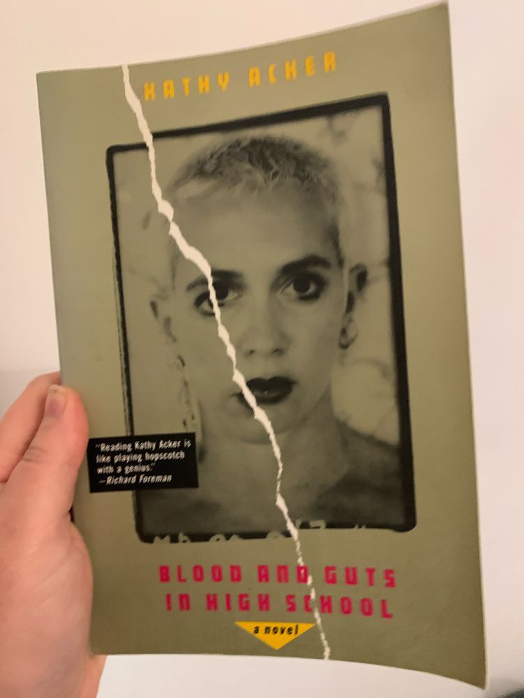 Book held up by a hand on the left side of frame. The boom has a greenish grey cover with the title in red sans serif text on the bottom that reads, “BLOOD AND GUTS IN HIGH SCHOOL” with “a novel” in lowercase black letters atop a small golden triangle and the author’s name “Kathy Acker” at the top of the cover in gold text. Cantered on the page is a photograph ripped in half of a white woman with closely shorn bleached hair and small ring earrings who looks blankly head on at the camera 