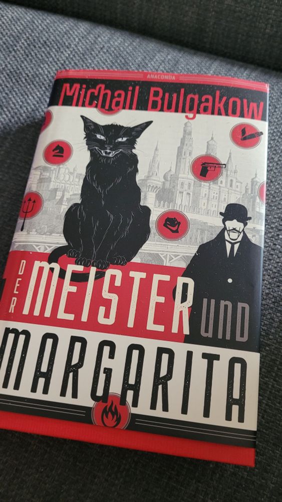 Das Buch "Der Meister und Margarita" von Michail Bulgakow. Ein großer schwarzer fauchender Kater und ein in Schwarz-Weiß gehaltener Mann im Mantel und Melone auf dem Kopf sowie Krawatte um den Hals. Im Hintergrund die Silhouette Moskaus sowie mehrere kleine in roten Kreisen eingefasste Symbole, z.B eine rauchende Zigarre, eine Pistole, ein Dreizack, ein Springer vom Schach und mehr.