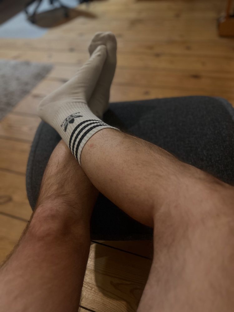 Two hairy legs, crossed and only wearing white Adidas Crew socks on a stool 
