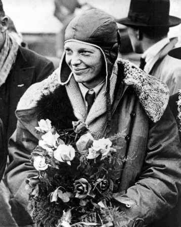 Amelia Earhart Celebrated After Her Transatlantic Solo Flight

In this historic black-and-white photograph, Amelia Earhart stands with a modest smile as she clutches a bouquet of roses. Clad in a heavy flight jacket, wool scarf, and her signature leather aviator cap, she is surrounded by well-wishers and onlookers, their faces turned toward the pioneering aviator in admiration. The image captures a moment of triumph shortly after Earhart’s groundbreaking solo transatlantic flight in May 1932—a daring 15-hour journey from Newfoundland, Canada, to a pasture in Northern Ireland. Her achievement was a milestone not only in aviation history but also in women’s rights, proving that women could match and surpass the feats of male aviators.

This photograph was likely taken during her return to the United States, where she was honored with widespread accolades and decorations. Most notably, on June 24, 1932, Earhart became the first woman awarded the Distinguished Flying Cross by the U.S. Congress, recognizing her courage and leadership in advancing aviation. At a time when women were expected to remain grounded—both figuratively and literally—Earhart took to the skies and rewrote the narrative. Her achievement made global headlines, inspiring generations of women to defy societal expectations and reach new heights.

The symbolic contrast between the flowers in her hand and the rugged aviation gear she wears reflects the dual image Earhart projected: grace paired with steel determination. This image thus not only commemorates a personal victory but also encapsulates a seismic cultural shift in public perception of women’s capabilities. Earhart didn’t just fly—she soared into history, earning her place not only in the sky but in the hearts of millions around the world.