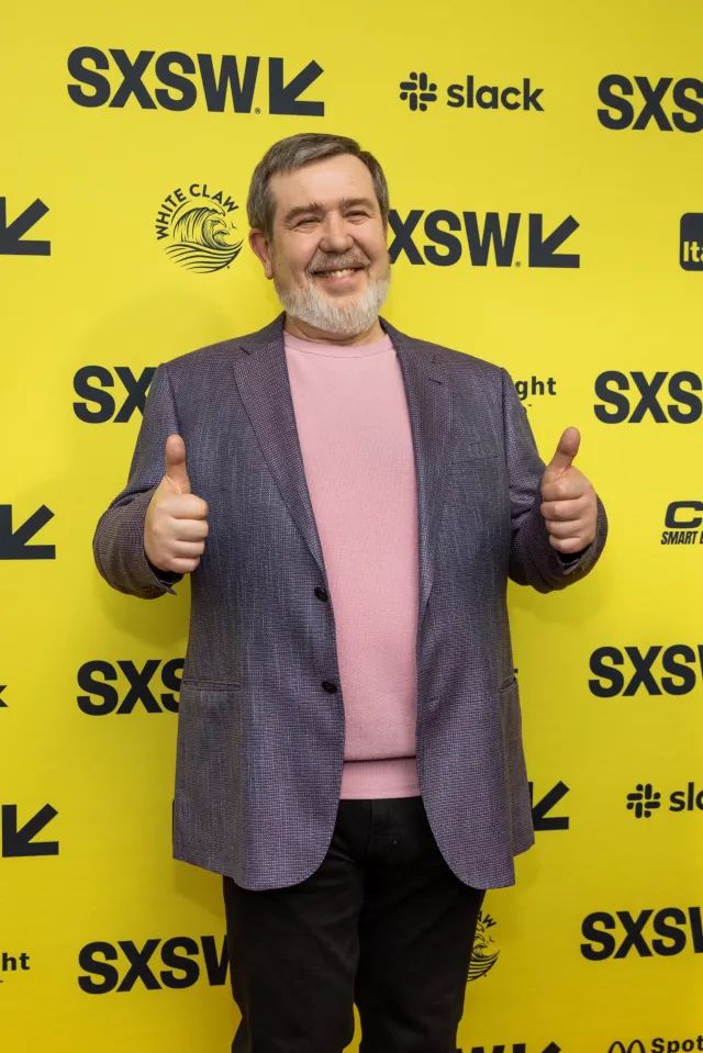 Alexey Pajitnov at SXSW – The Enduring Legacy of Tetris

In this modern-day photograph, Alexey Pajitnov—creator of Tetris—stands beaming with both thumbs up, captured at the SXSW (South by Southwest) festival in front of a bright yellow step-and-repeat wall. Clad in a lavender blazer over a soft pink sweater, Pajitnov appears relaxed, jovial, and warmly received by an audience that has grown up with his creation. While the backdrop features logos of global tech and media brands like Slack and White Claw, the real spotlight here is on Pajitnov himself, whose enduring legacy stretches across generations and continents. His attendance at SXSW underscores the remarkable journey of Tetris—a game that began as a humble side project in a Moscow computing lab in 1984 and went on to reshape the global gaming landscape. Unlike many developers of that era, Pajitnov saw his creation become a cultural phenomenon not just once, but repeatedly: from the arcade boom to Game Boy success, mobile gaming revolutions, and now modern reimaginings in film and augmented reality. His smile speaks to the quiet pride of a man who bridged East and West not through politics, but pixels. This image not only honors his contribution to game design but also celebrates how intellectual curiosity and elegant design can create tools of joy and unity. At SXSW—a festival that celebrates boundary-breaking creativity—Pajitnov is not just a nostalgic figure. He’s a living reminder that some of the most transformative innovations come from the unlikeliest of places and continue to evolve long after their inception. From Cold War obscurity to pop culture icon, this photo affirms Pajitnov’s place among the digital age’s great innovators, as a symbol of playful resilience and the global language of games.