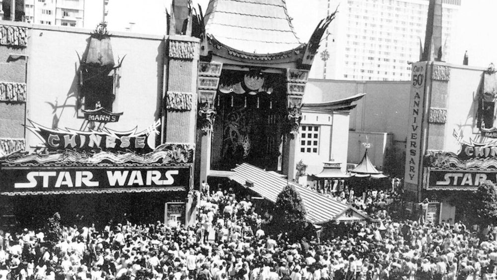 The Birth of a Blockbuster – Star Wars Premieres at Mann’s Chinese Theatre, 1977

This iconic black-and-white photograph captures the explosive premiere of Star Wars on May 25, 1977, outside Mann’s Chinese Theatre in Hollywood, California. The building’s distinct East Asian architectural style forms a dramatic backdrop, with its ornate rooflines and pagoda-style details, providing a fittingly theatrical setting for a moment that would redefine the moviegoing experience forever. Beneath the towering marquee boldly emblazoned with “STAR WARS,” an enormous crowd stretches across the sidewalk and beyond, emphasizing the instant magnetism of the film. At the time, Star Wars had only opened in 32 theaters across the United States, but word-of-mouth, combined with unprecedented enthusiasm, sparked long lines and sold-out shows within hours. What’s seen here is not just a queue—it is a cultural ignition point. The crowd represents a cross-section of curious sci-fi fans, movie lovers, and pop culture enthusiasts, many of whom had no idea they were witnessing the dawn of a cinematic revolution. George Lucas’s vision had blended mythological themes, space opera grandeur, and pioneering visual effects into a genre-defining masterpiece. The audience outside Mann’s Chinese Theatre wasn’t merely waiting to see a movie—they were stepping into a mythos that would soon expand into a galaxy-spanning franchise, inspire generations of creators, and shift the Hollywood model toward blockbuster filmmaking. The photo freezes a transformative day, where cinema history and global pop culture converged under a single marquee.