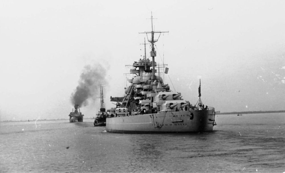The Bismarck Sets Sail — Prelude to the Hunt

This historic black-and-white photograph captures the German battleship Bismarck as it departs from port on May 19, 1941, launching one of the most infamous naval operations of World War II. Towering at the center of the frame, the Bismarck exudes sheer military might, with its heavily armored hull, massive primary gun turrets, and complex radar and communications rigging prominently visible. Tugboats assist her movement, while a plume of smoke rises from a support ship in the background, symbolizing the tense departure into open waters. This image encapsulates a moment just before history would take a dramatic turn. At over 50,000 tons and equipped with 15-inch guns, the Bismarck was designed as a strategic weapon to disrupt Allied supply lines in the Atlantic. Her breakout from Gotenhafen into the North Sea—code-named Operation Rheinübung—would trigger a relentless pursuit by the Royal Navy. Less than ten days later, the ship would be hunted down and sunk after destroying HMS Hood, the pride of the British fleet. The image evokes not just the engineering prowess of Nazi Germany, but also the fragile supremacy of battleship warfare in an era increasingly defined by air power and radar-guided detection. The serene waters surrounding the vessel starkly contrast with the chaos and destruction that would soon follow. This departure marked the beginning of the Bismarck’s short but devastating legacy, which would culminate in her destruction on May 27, 1941—a symbolic victory for the British and a sobering reminder of the brutal chessboard of WWII naval conflict.