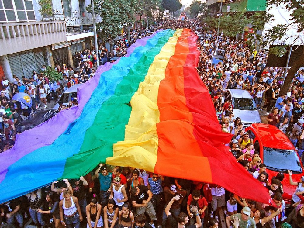 A Sea of Pride — Marching Beneath the Rainbow Banner

This vivid and electrifying photograph captures the exuberant spirit of a modern Pride parade, with thousands of people marching beneath a massive, undulating rainbow flag that stretches the entire width of a city street. Shot from above, the image offers a striking visual of coordinated community movement—an expression of collective visibility and joy. The rainbow fabric flows like a river through the urban canyon, its vibrant colors glowing in the sunlight. Red, orange, yellow, green, blue, and purple ripple in rhythm with the crowd’s energy, reinforcing the symbolic unity of diversity that the flag has come to represent. Participants of all ages, races, and gender identities walk together in solidarity, some carrying the flag, others raising their arms in celebration or snapping photos of the moment. Above them, apartment buildings and balconies are lined with onlookers and allies, reinforcing that this is not a fringe celebration—it is deeply rooted in the heart of the city.

This moment, while celebratory, is the culmination of decades of struggle. The flag they carry is the legacy of Gilbert Baker’s original 1978 design, now universally recognized as the symbol of LGBTQ+ pride and resilience. Each color represents more than a hue; it conveys an aspect of human experience and queer identity: life, healing, sunlight, nature, harmony, and spirit. In recent decades, the Pride flag has evolved to be even more inclusive, incorporating colors representing trans, nonbinary, intersex, and BIPOC communities—reminding us that the journey for justice is still ongoing. This image encapsulates both that progress and that purpose. It is not just a celebration of identity; it’s a living act of resistance and remembrance. The street becomes a stage, the flag a banner of defiance, love, and visibility, moving forward with every step, every beat, and every raised voice under its colors.