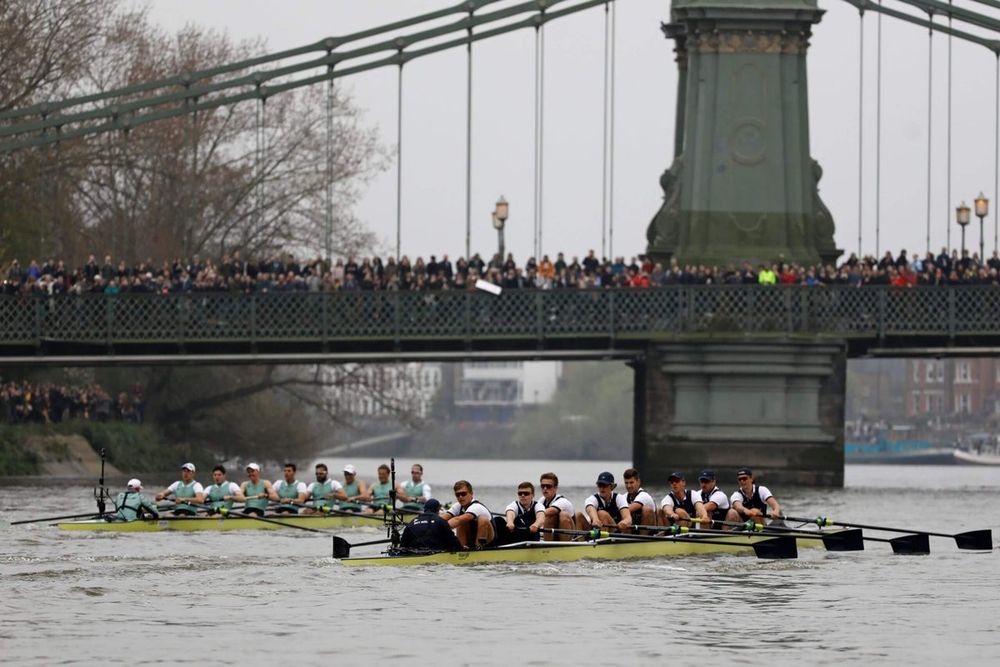 A Tradition in Motion – Modern Oxford-Cambridge Boat Race on the Thames

This captures the Oxford and Cambridge rowing crews in the midst of a modern-day Boat Race on the River Thames, passing beneath the Hammersmith Bridge. Both boats, sleek and brightly painted in their traditional colors—dark blue for Oxford, light blue for Cambridge—are in fierce competition, rowers in matching uniforms pulling in perfect synchronization. On the bridge and along the riverbanks, large crowds of spectators gather to watch, signaling the enduring popularity of this storied event. Smartphones and modern attire among the crowd contrast with the timelessness of the race itself, merging history with the present.

Held annually since 1829, the Oxford-Cambridge Boat Race has evolved from a gentleman’s challenge into a global sporting spectacle. This modern image reflects advancements in both athletic preparation and broadcasting. Today, teams train year-round with sports physiologists, nutritionists, and Olympic-level coaches. Sophisticated GPS tracking, drone footage, and real-time analytics now accompany each race, bringing new dimensions to strategy and storytelling. The introduction of women’s and lightweight races in recent decades has also modernized the tradition, expanding its inclusivity and reach.

The photo captures a pivotal moment—every stroke is an assertion of legacy, pride, and institutional rivalry. Each crew races not just to win, but to etch their names into the long annals of Boat Race history. The symmetry of the rowers, the focus in their eyes, and the crowd’s collective anticipation create a tableau of high athletic drama. Hammersmith Bridge, itself an emblem of Victorian engineering and London’s urban identity, looms as a silent witness to this clash of elite endurance.

This underscores how some traditions, even as they modernize, retain their soul. The Oxford-Cambridge Boat Race continues to embody competition, camaraderie, and the unbroken spirit of tradition.