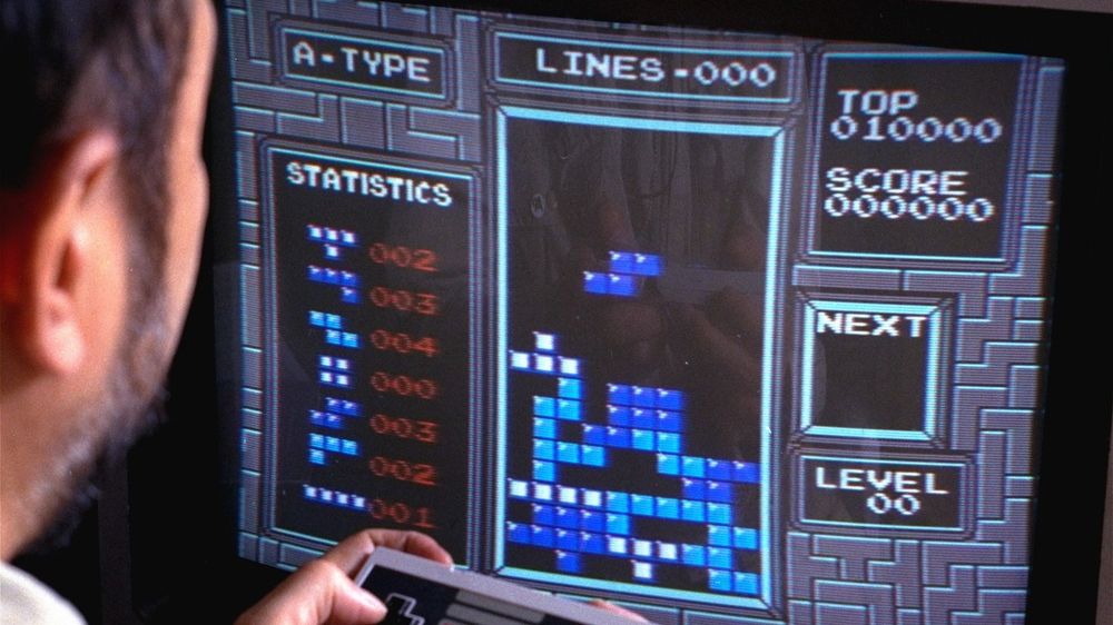 Playing Tetris on NES – The 8-Bit Global Invasion

This image captures the unmistakable sight of Tetris in action on a Nintendo Entertainment System (NES), the platform that helped solidify the game’s legendary status outside the Soviet Union. A player—seen from behind—is holding a rectangular NES controller, focused intently on the screen as tetrominoes descend. The familiar blue-on-black grid showcases the simplicity and addictiveness that made Tetris a transcendent hit. To the right, the score and level indicators are blank, suggesting the beginning of a game. On the left, statistical readouts document the frequency of each tetromino’s appearance, adding a layer of depth for players obsessed with optimizing their moves. The screen is washed in the glowing blur of CRT scan lines, transporting the viewer back to a time when 8-bit aesthetics were the cutting edge of home entertainment.

While Tetris was originally developed on a Soviet computer in 1984, it was its release on the Game Boy and NES in the late 1980s that launched the game into global superstardom. Henk Rogers, a Dutch-American entrepreneur, played a crucial role in negotiating the Western publishing rights and partnering with Nintendo to bundle the game with its new handheld console. This strategic move didn’t just sell millions of Game Boys—it turned Tetris into a symbol of cross-cultural exchange in the waning years of the Cold War. Despite geopolitical divisions, this small puzzle game became a common language among gamers across the globe.

This image, though simple at first glance, represents how an abstract Soviet algorithm made its way into living rooms from Tokyo to Toronto. Here, as tetrominoes fall and lines clear, history itself plays out in silent increments. It’s a reminder that some of the most enduring cultural legacies don’t come from grand speeches or world leaders—but from clever code, falling blocks, and a well-timed rotation.