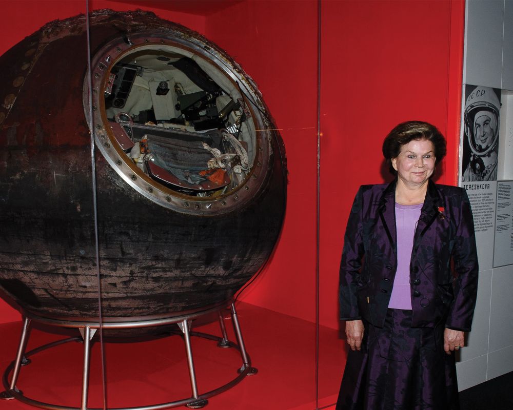 Legacy and Artifact – Valentina Tereshkova and the Vostok 6 Capsule

In this photograph, Valentina Tereshkova, now an older woman and decorated veteran of space history, stands proudly beside the very spacecraft that made her a global icon: the Vostok 6 descent module. Encased in a museum display and scorched black from atmospheric reentry, the spherical capsule bears the wear of one of the most historic journeys in space exploration. Its compact interior is visible through the open hatch, revealing the cramped, utilitarian design of early Soviet spacecraft. Tereshkova, dressed in a formal plum-colored suit, smiles gently—embodying both dignity and quiet pride—as she poses before the vessel that launched her into history on June 16, 1963.

This moment is more than a reunion with an artifact; it is a meeting of legacy and memory. The Vostok 6 capsule represents a pivotal era when the Soviet Union competed fiercely in the Space Race, and Tereshkova’s mission was a major geopolitical and cultural milestone. She became not only the first woman in space, but also the only woman to fly solo in orbit—a feat unmatched to this day. Her mission challenged entrenched gender roles and sparked global conversations about women’s roles in science and engineering.

The juxtaposition of Tereshkova and the capsule decades after the flight emphasizes the endurance of her legacy. It reflects how pioneers in space exploration are not merely participants in scientific milestones but also powerful symbols of human potential, courage, and change. The exhibit surrounding her includes a poster with her flight portrait and name in Cyrillic script, underscoring her continued reverence in Russian memory and global history. Today, her flight is remembered not just as a Cold War achievement, but as a human story of aspiration, resilience, and the determination to reach beyond what had previously been deemed possible.