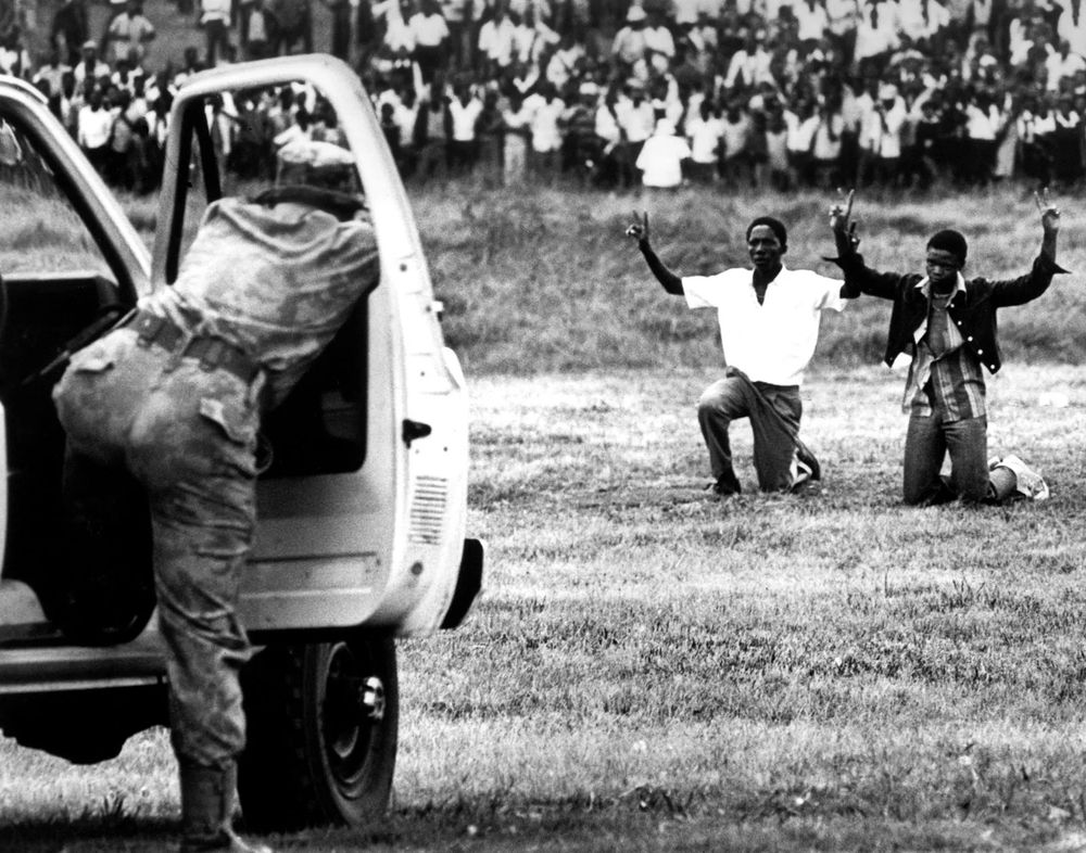 Hands Raised in Defiance – Confronting Apartheid Power

This striking black-and-white photograph captures a tense and heartbreaking moment from the Soweto Uprising. Two young Black South African men are seen kneeling on the grass with their hands raised in surrender, directly facing a white South African security officer who is leaning out of a vehicle, weapon aimed. The young men’s expressions are unreadable from the photo’s angle, but their posture—arms lifted in submission, knees grounded—is a haunting depiction of vulnerability under authoritarian force. Behind them, a crowd of onlookers looms in the background, their presence simultaneously witness and silent protest. This image speaks volumes about the brutal power imbalance that characterized apartheid South Africa. The act of raising hands, a universal gesture of “don’t shoot,” becomes here a chilling symbol of systemic violence and the cost of peaceful protest. Taken during the escalation of demonstrations that followed the June 16, 1976 massacre, including events on June 20, it highlights the militarized response of the apartheid regime to youth-led demands for justice and education. The uniformed soldier, partially shielded by the vehicle’s open door, represents a faceless state apparatus—mechanized, impersonal, and lethal. The stark contrast between the young men’s simple clothing and the soldier’s gear emphasizes the imbalance of force. This photograph encapsulates not only the terrifying risks protestors faced, but also their defiant courage. It serves as a historical reminder of how movements are met with resistance—and how even unarmed voices can rise to confront institutionalized oppression. The image is both a document of injustice and a testament to resilience.