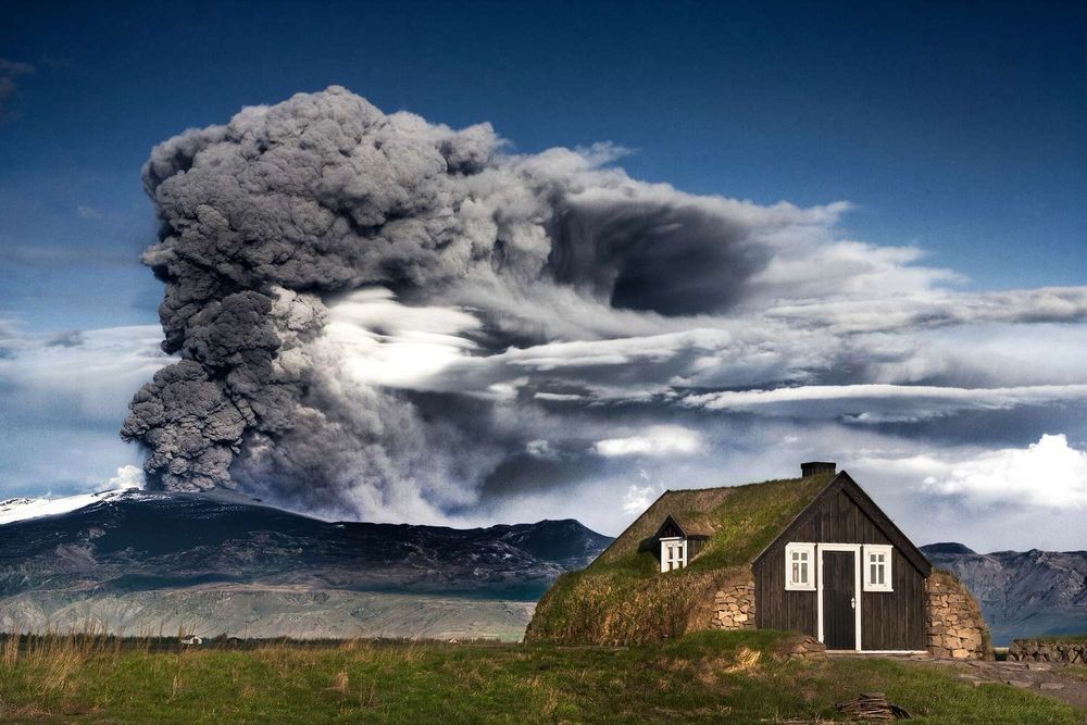 Eyjafjallajökull’s Ash Tower Looms Over Rural Iceland – Nature’s Spectacle Meets Daily Life

This striking image captures a towering ash plume erupting from Iceland’s Eyjafjallajökull volcano during its 2010 eruption, seen from a tranquil rural farmstead in the Icelandic countryside. The contrast between the serene, turf-roofed black wooden cottage in the foreground and the billowing, mushrooming cloud of ash and volcanic gases in the background creates a surreal visual dichotomy between human habitation and the raw power of nature. The Eyjafjallajökull eruption lasted 39 days, beginning in April and officially ending on May 22, 2010. During that time, it spewed an estimated 250 million cubic meters of volcanic material into the atmosphere. The massive ash cloud, lofted high into the jet stream, grounded much of Europe’s air traffic for nearly a week and impacted more than 10 million travelers, highlighting how profoundly natural events can disrupt global infrastructure. Yet for many Icelanders living near the eruption, life carried on under the ever-present shadow of volcanic ash. Iceland’s history is intimately tied to its geological volatility, and this photo captures that enduring coexistence. The turf-roofed house reflects centuries-old Icelandic architectural traditions that prioritized insulation and harmony with the land, making it both a symbol of cultural resilience and sustainable living. As Eyjafjallajökull sent dark clouds into the stratosphere, disrupting economies and headlines worldwide, the people living nearby continued their routines, adapting once again to the island’s fiery temperament. This image serves as a powerful reminder of humanity’s small yet enduring presence in the face of Earth’s elemental forces.