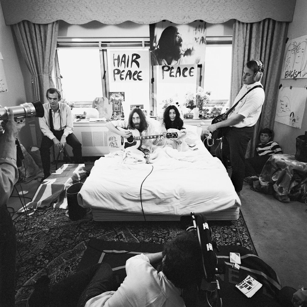 A Song for Peace – “Give Peace a Chance” is Born

This evocative black-and-white image captures the creative and cultural climax of John Lennon and Yoko Ono’s second “Bed-In for Peace,” which began on May 26, 1969, in Montreal. The photograph shows the couple sitting upright in bed surrounded by microphones, audio cables, and musical instruments as they record the now-iconic antiwar anthem “Give Peace a Chance.” Lennon, with his signature long hair and round glasses, strums an acoustic guitar, while Yoko Ono sits beside him in serene stillness, symbolizing both partnership and protest. Around them, a small group of musicians, sound engineers, and journalists crowd into the modest hotel room, which has been transformed into an unlikely recording studio. Signs above their heads read “HAIR PEACE” and “PEACE,” echoing the couple’s core message and acting as backdrops to one of the most memorable acts of musical activism of the 20th century. The atmosphere in the room blends intimacy with urgency, making the recording session feel like both a jam session and a political demonstration. This image immortalizes the moment when the ideals of the 1960s counterculture—peace, resistance, and global unity—were distilled into a single phrase repeated in chorus. The song, improvised and recorded with minimal equipment, became the rallying cry of antiwar protests around the world, used in demonstrations from the Vietnam War era to more recent movements. It marked the moment when pop culture and political dissent fused through performance art, creating an enduring global symbol of peaceful resistance. More than a publicity stunt, the Bed-In became a platform where celebrity, creativity, and conviction converged to challenge violence with music, humor, and radical vulnerability. In this image, we witness the transformation of a bedroom into a battlefield of ideas—one fought not with weapons, but with chords and convictions.