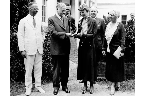 Amelia Earhart Receives the Distinguished Flying Cross from President Herbert Hoover

This historic black-and-white photograph captures a defining moment in Amelia Earhart’s storied career: the ceremonial presentation of the Distinguished Flying Cross by President Herbert Hoover on June 24, 1932. The scene is set outdoors on the White House lawn, with five individuals forming a semicircle of recognition around the famed aviator. At center, President Hoover, dressed in a dark suit, leans forward to present the medal to Earhart, who stands with quiet dignity, eyes lowered, and hands gently clasped to receive the honor. Her simple yet elegant attire—a tailored, double-breasted dress with buttons running down the front—underscores her practical nature and unassuming style, even as she makes history.

Surrounding them are Vice President Charles Curtis and First Lady Lou Hoover, joined by other dignitaries and aviation leaders, all bearing witness to this symbolic moment. The event marked the first time the Distinguished Flying Cross—a military decoration usually reserved for valor or achievement in aerial flight—was awarded to a woman. Earhart earned this recognition after completing her daring solo transatlantic flight on May 20–21, 1932, becoming the first woman to cross the Atlantic alone by air. Her flight not only mirrored Charles Lindbergh’s solo achievement five years earlier but surpassed it in difficulty due to poor weather and mechanical troubles.

This image speaks volumes about changing societal tides. Earhart’s presence at the White House was more than ceremonial—it was a statement of progress. In an era when female pilots were still rare and often doubted, Earhart shattered gender barriers with quiet confidence and unmatched courage. This moment helped cement her role as both an aviation pioneer and a cultural icon whose legacy would continue to inspire future generations of women in science, exploration, and leadership.