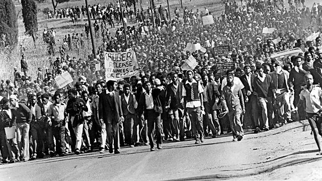 A Tide of Resistance – The March of Soweto’s Youth

This powerful black-and-white photograph captures an immense procession of Black South African students and youth marching in unison during the Soweto Uprising of June 1976. Taken from a frontal angle, the image shows thousands of demonstrators moving resolutely down a wide street, their faces serious, focused, and unafraid. In the front row, several young men walk shoulder to shoulder, leading the march with signs held high. One placard pleads: “KRUGER PLEASE DETAINEES CRIES,” referencing South African Minister of Police Jimmy Kruger and the brutal detainment and torture of students following the June 16 massacre. The scale of the demonstration is staggering; the crowd stretches into the distant horizon, creating a wave-like impression of relentless determination. This march likely took place on or around June 20, during the sustained protests that followed the initial killings by police. What began as a school walkout against the forced instruction of Afrikaans rapidly became a nationwide rebellion against the entire apartheid system. The orderly defiance of this image contrasts starkly with the violence these students would soon face. There are no weapons here—only resolve. The photograph evokes the spirit of youth-led civil disobedience, echoing movements around the world yet grounded in the specific brutality of apartheid. Every face in the crowd is part of history: children turned freedom fighters, armed only with voices, signs, and the will to be heard. This image embodies the raw courage and unified resistance that turned Soweto into a symbol of revolution and lit a fire that would burn through the decades until apartheid’s end. It is a visual record of humanity’s fight for dignity, one step at a time.